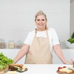 A mature woman in an apron stands at a kitchen counter with fresh produce and cheese, ready for cooking.
