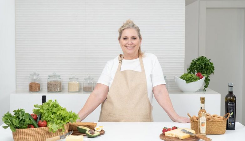 A mature woman in an apron stands at a kitchen counter with fresh produce and cheese, ready for cooking.